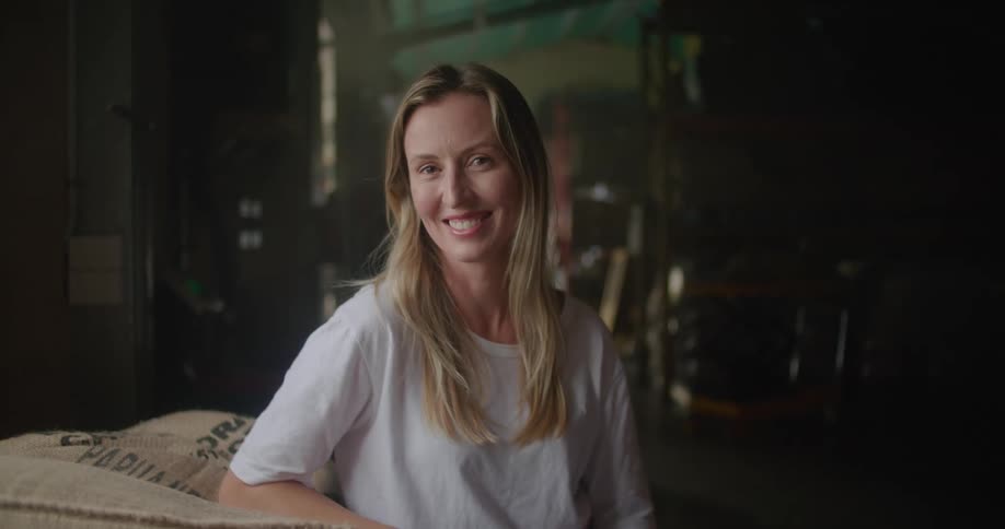 Blonde woman with white t shirt leans against coffee sacks looking and camera and smiling in coffee warehouse