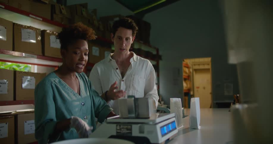 Man with white shirt and woman with curly hair earring and light blue shirt talk to each other measuring coffee beans in front of the shelves in coffee warehouse