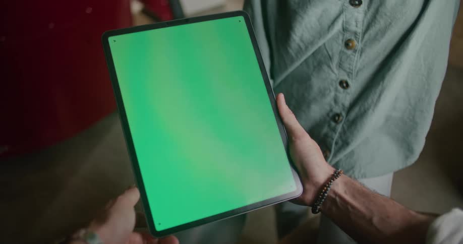Man with grey shirt and bracelet holds tablet with solid green screen indoors