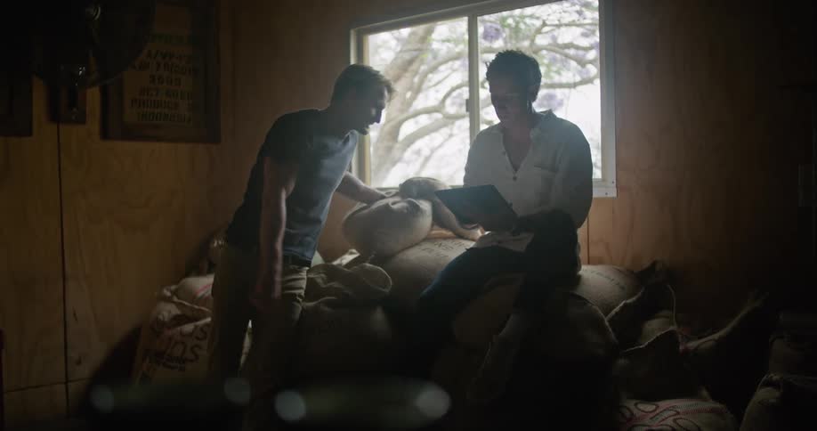 Man with white shirt sitting on coffee sacks uses tablet and talks to blonde man with dark blue shirt and brown pants in coffee warehouse