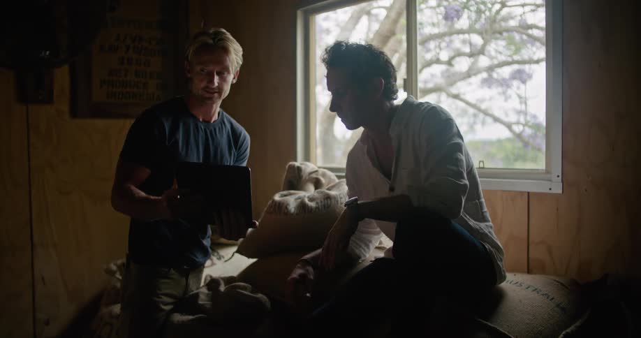 A man sitting on a pile of burlap sacks working on a tablet hands it over to a coworker as they point and talk