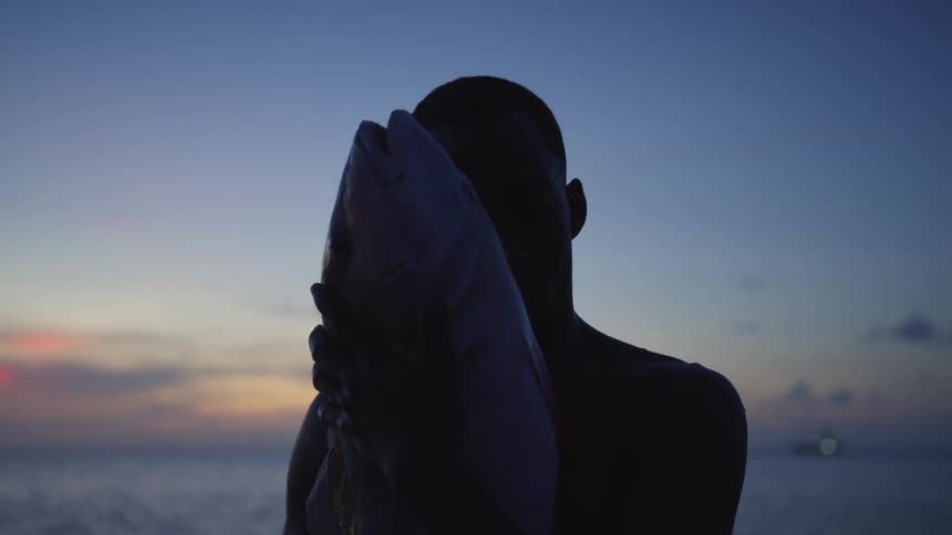 Silhouette of a man standing near the water holding a large fish in his hands as the sun sets on the horizon
