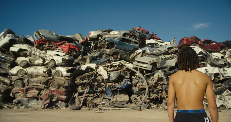 Youth stand topless staring at hill of scrapped cars