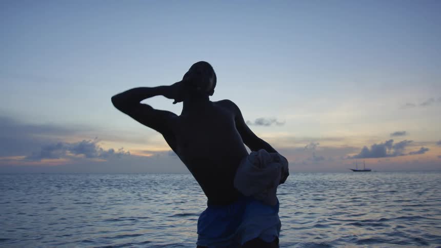 Young shirtless man dancing with a piece of clothing in his hands on the ocean at sunset