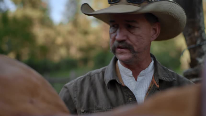 A man in a sunhat inspects the saddle on his horse during a break of their trip in the forest