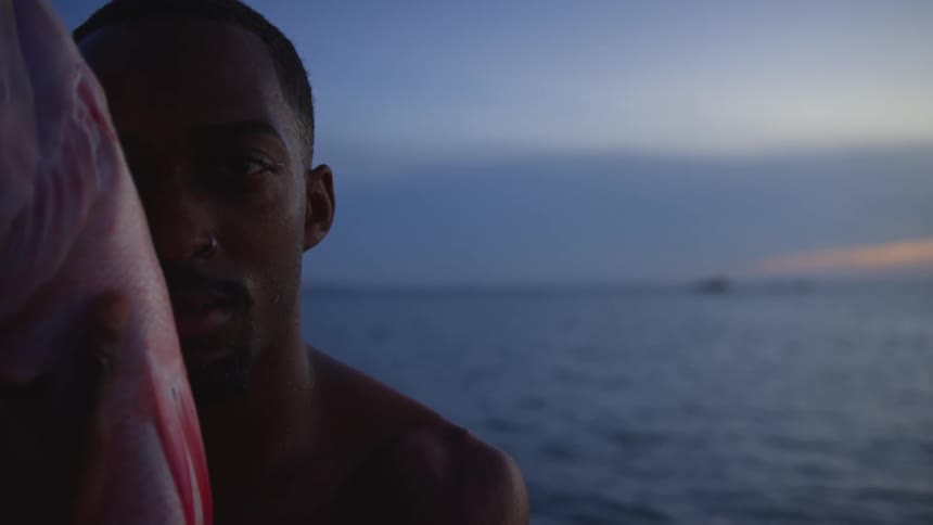 Shirtless man holding a dead fish close to his face in front of the ocean at sunset