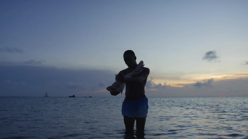 A man stands in the ocean at dusk wiping his chest with a towel or shirt