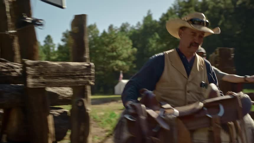 Cowboy enters a corral through a gate in the wooden fence carrying a horse saddle