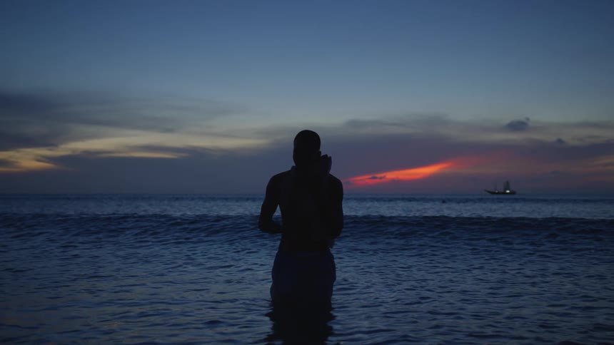 Man stands in ocean water silhouetted by crimson sky with ship on the horizon in the background