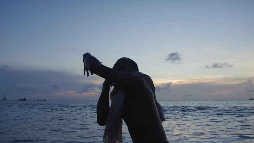 Shirtless man with towel around neck dancing is silhouetted against the sky on the beach at dusk