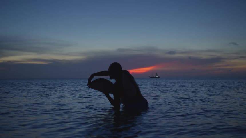 Silhouette of a man standing in the water holding a large fish in his hand as the sun sets on the horizon