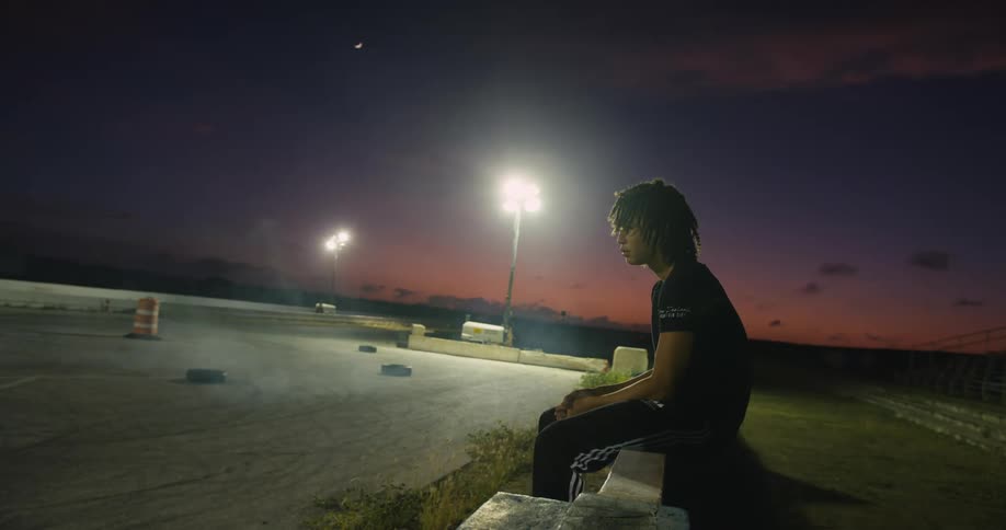 Young man sitting on a wall near an emtpy lot with blowing white smoke at night