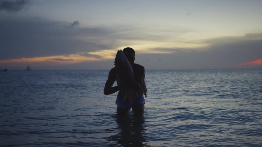 Silhouette of a man standing in the water holding a large fish in his hands as the sun sets on the horizon