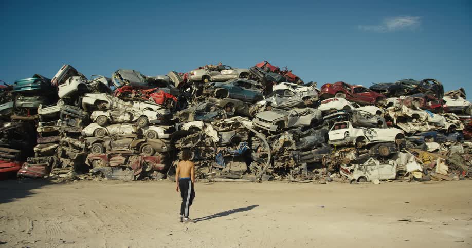 Topless youth walks back and forth on dirt looking at large pile of wrecked cars