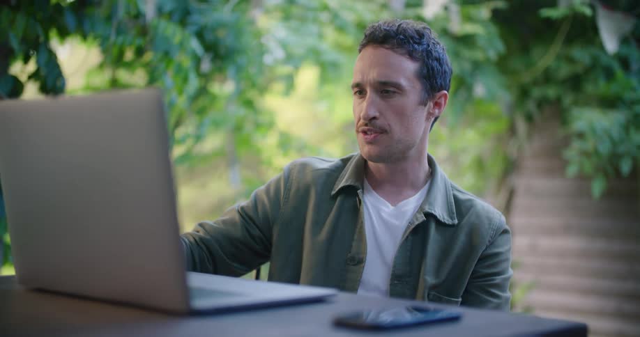 A man drinks a glass of water while participating in a video chat on a laptop at an outdoor table during the day