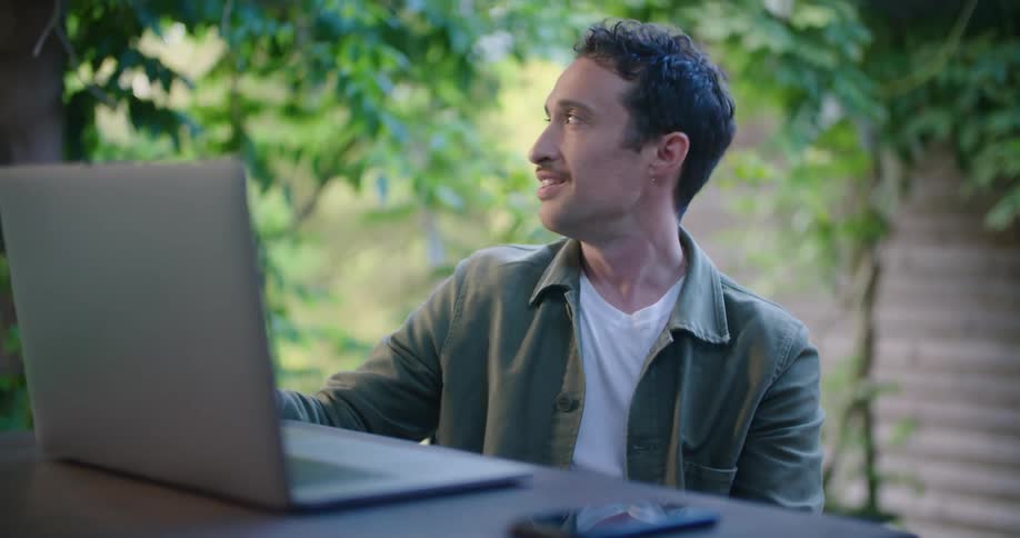 A man uses a laptop to participate in a video chat while sitting at a table outdoors during the day