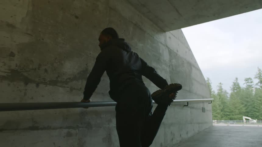 A man in athletic wear stretches his leg while leaning on a handrail in a concrete corridor next to an athletics field during the day