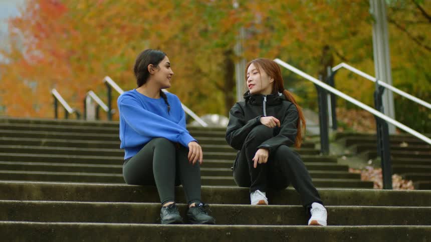 Woman joins friend on steps then they talk near trees with fall foliage