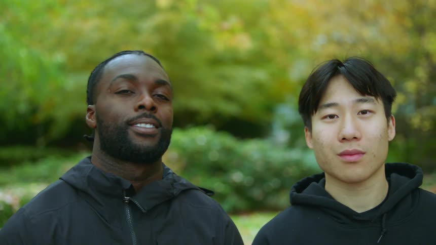 Two men wearing black hoodie and raincoat stare at camera smiling and laughing in green public park