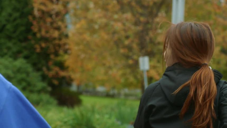 Woman with pony tail and black raincoat jogs with woman wearing blue sweater and man with braids and black rain coat in public park with autumn trees