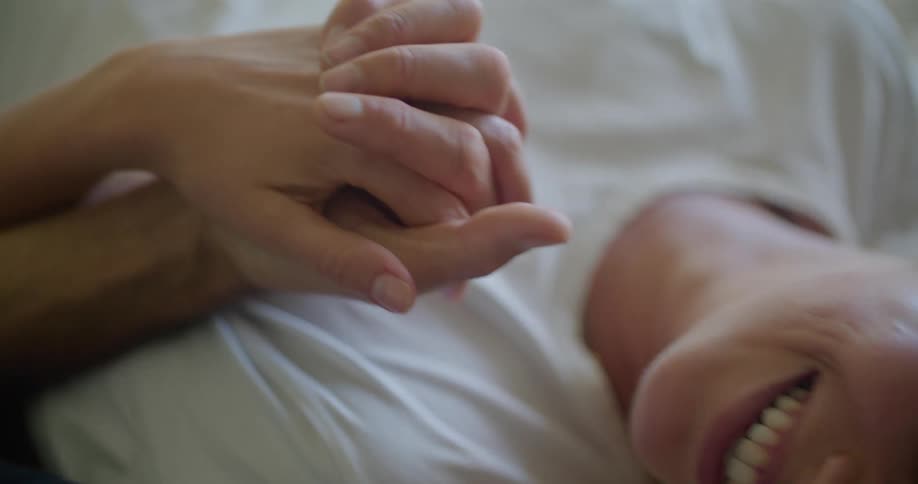 Couple lie on bed together holding hands then smile before kissing
