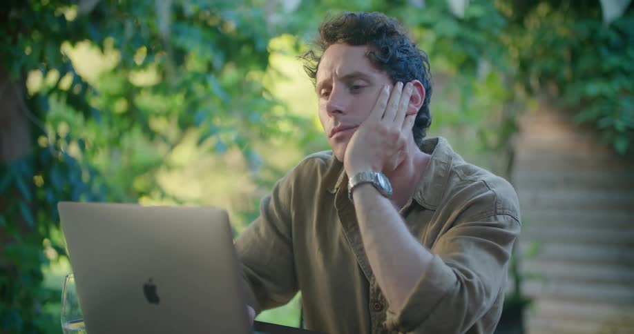Man with green shirt and wristwatch sits at table and thinks before typing on laptop in green garden