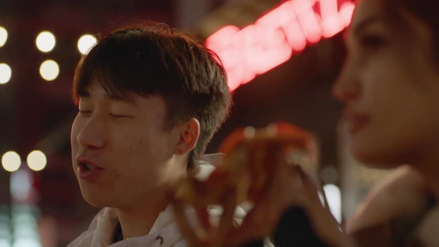 Friends sit outdoors near neon signed building eating pizza