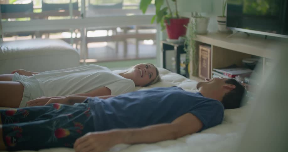 A couple sitting on a mattress fall back at the same time while looking at each other