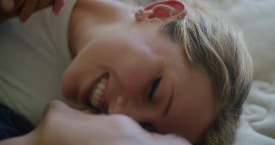 Couple lying on white quilt smiles and kisses while holding hands