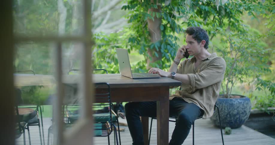 Young man sitting at a table on the porch talks on the phone while looking at a laptop