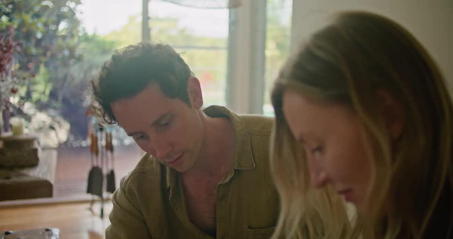 Man with yellow shirt and black pants and blonde woman wearing black blouse sit on beige couch working on documents talking to each other in living room at home