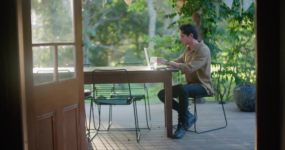 Man sits on a talbe on the porch to use a laptop while petting his dog