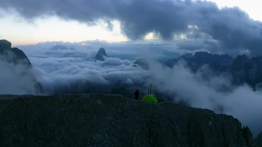 Aerial view of a green tent in the peak of a mountain range covered in clouds at sunset
