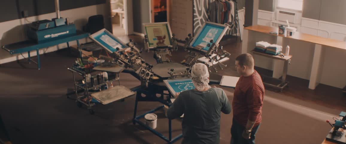 Man joins another man using silk screening machine in workshop