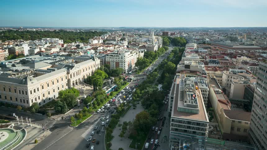 Traffic flows along streets through low lying buildings than people stand near building with imaginative architecture