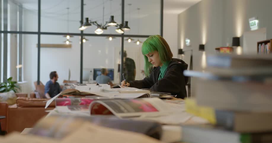 Girl with green hair writes at counter as men in glassed in office view screen