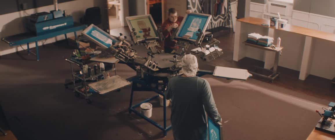 Two workers use silk screening machine in printing company workshop