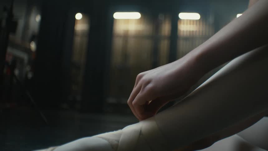 A ballet dancer sits on the floor and tightens the straps of her pointe shoes