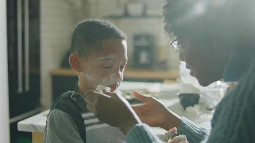 A woman cleans the flour off a boys face by blowing on it and wiping it with a napkin before giving the boy a kiss