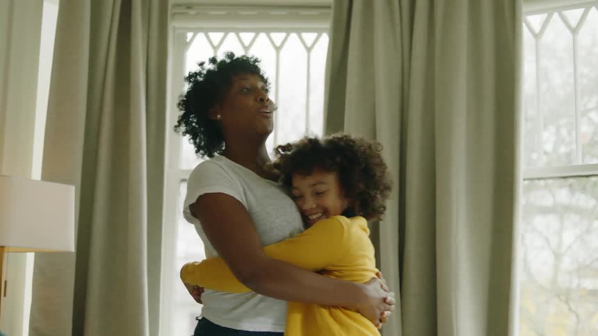 A woman hugs her daughter in the living room of a house as her husband and son sit on a nearby sofa