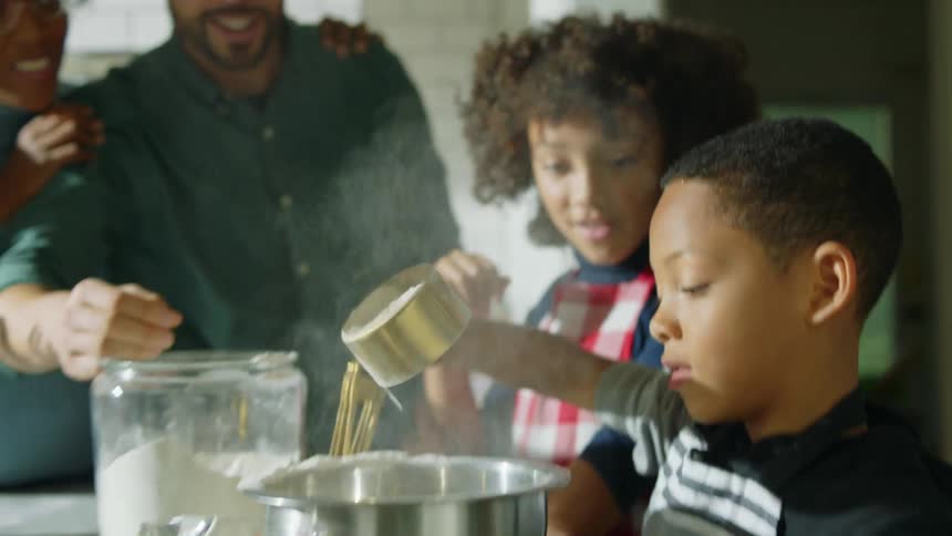 A boy sifts flour into a mixing bowl as his family watches from his side