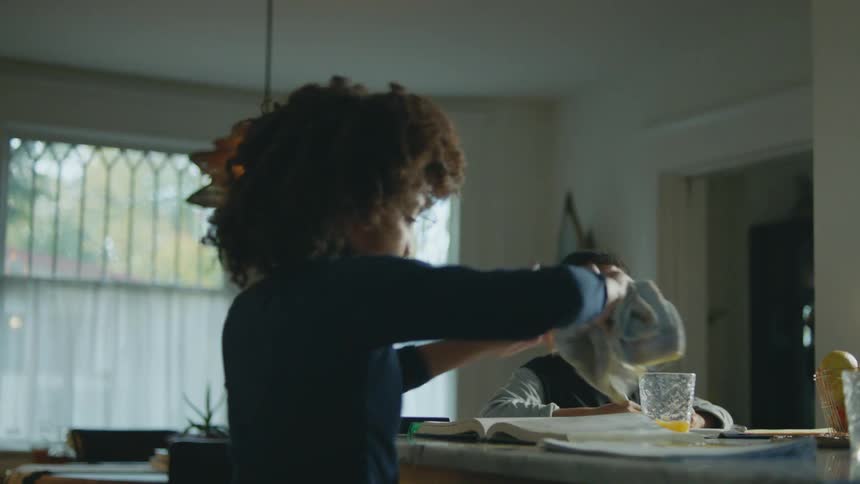 A girl spills orange juice on her homework causing her mother to rush off to help her clean it up