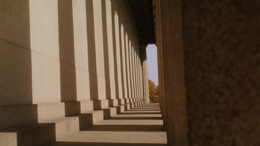 Sunlight through tall columns casts shadows in the outdoor corridor of a large building