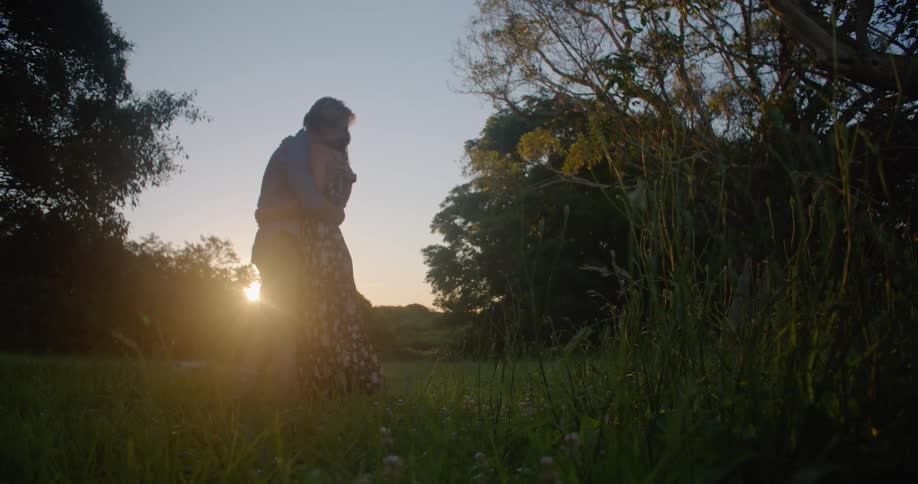A man and a woman embrace and kiss and her swings her around as the sun shines brightly behind them