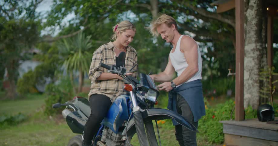 A woman sits on the motorcycle with her phone as a man repairs it