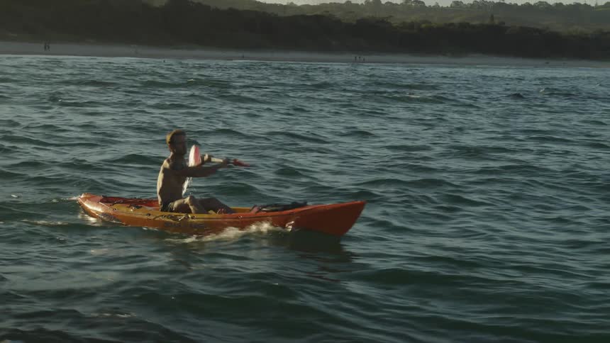 A man paddles a canoe over waves in a lake as people in the background walk on the beach