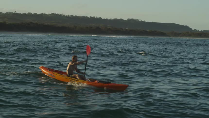 Shirtless young man paddling on a kayak on the ocean near the coast at sunset