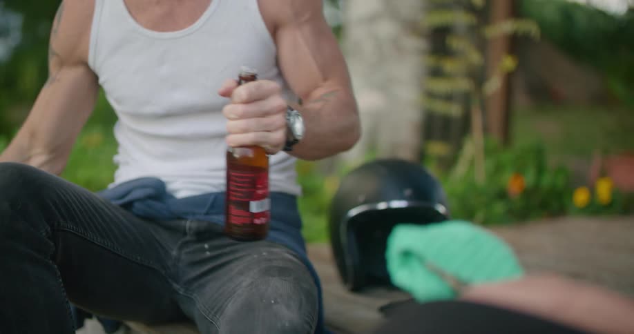 A man sitting outdoors uses a bottle opener to open a bottle of beer before tapping it against the bottle of a person sitting across from him