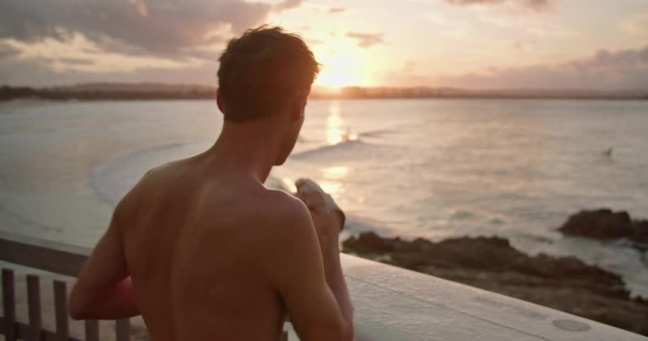 A man stands on a balcony on the shore and takes his shirt off and sets it on his surfboard as he looks out at the water