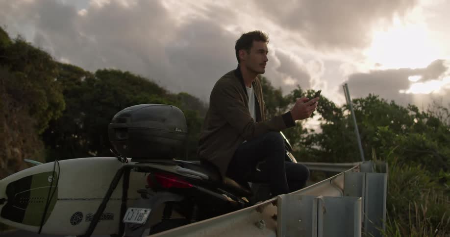 Young man sitting on a motorcycle looking at his phone at sunset